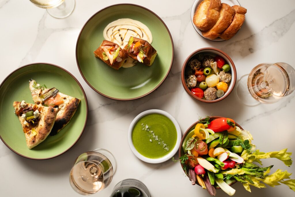 Colorful spread of gourmet dishes, including flatbread, lobster rolls, vegetables, and wine
