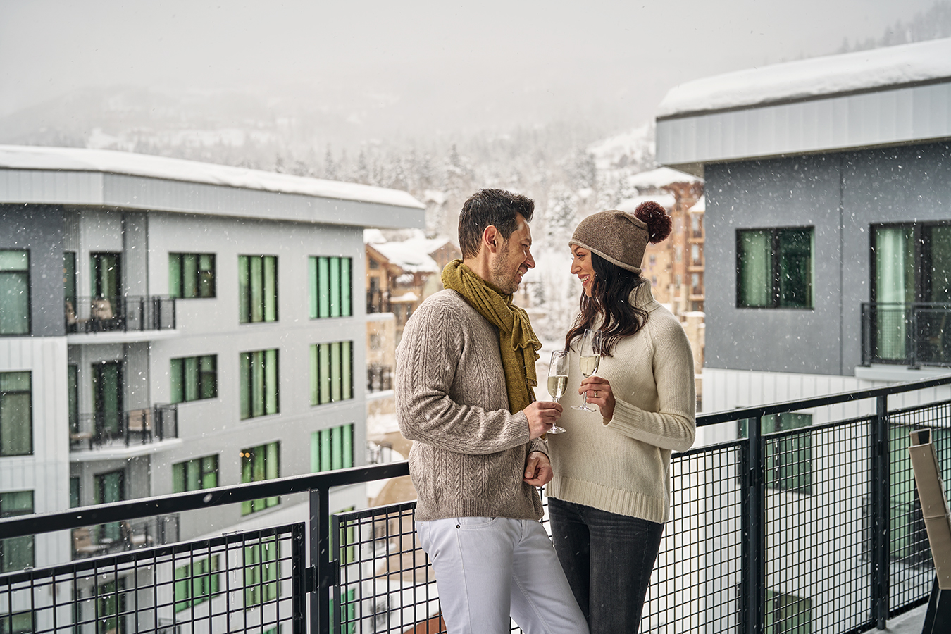 A couple dressed in cozy winter sweaters enjoys champagne together on a snowy balcony, surrounded by modern mountain lodges and snow-covered trees
