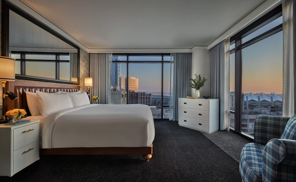 Hotel suite with a large bed, city views, and floor-to-ceiling windows at sunset