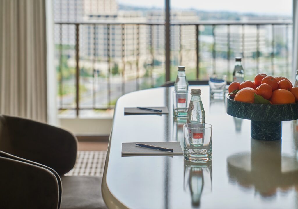 Close-up of a meeting table with bottled water, notepads, pens, and a bowl of oranges, with a city view in the background