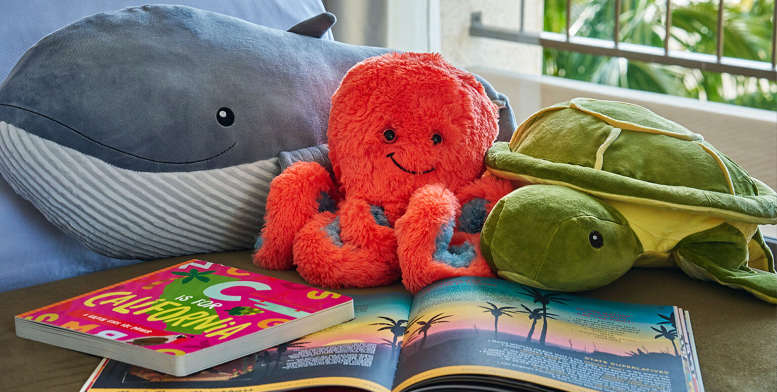 Three stuffed animals sitting on a couch next to children's books at Pendry Newport Beach