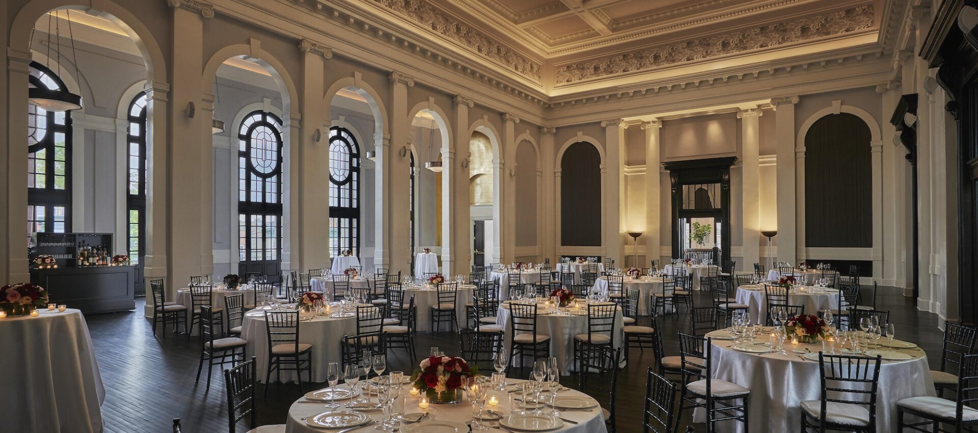 Grand ballroom with high ceilings, arched windows, round tables, and elegant dinner settings