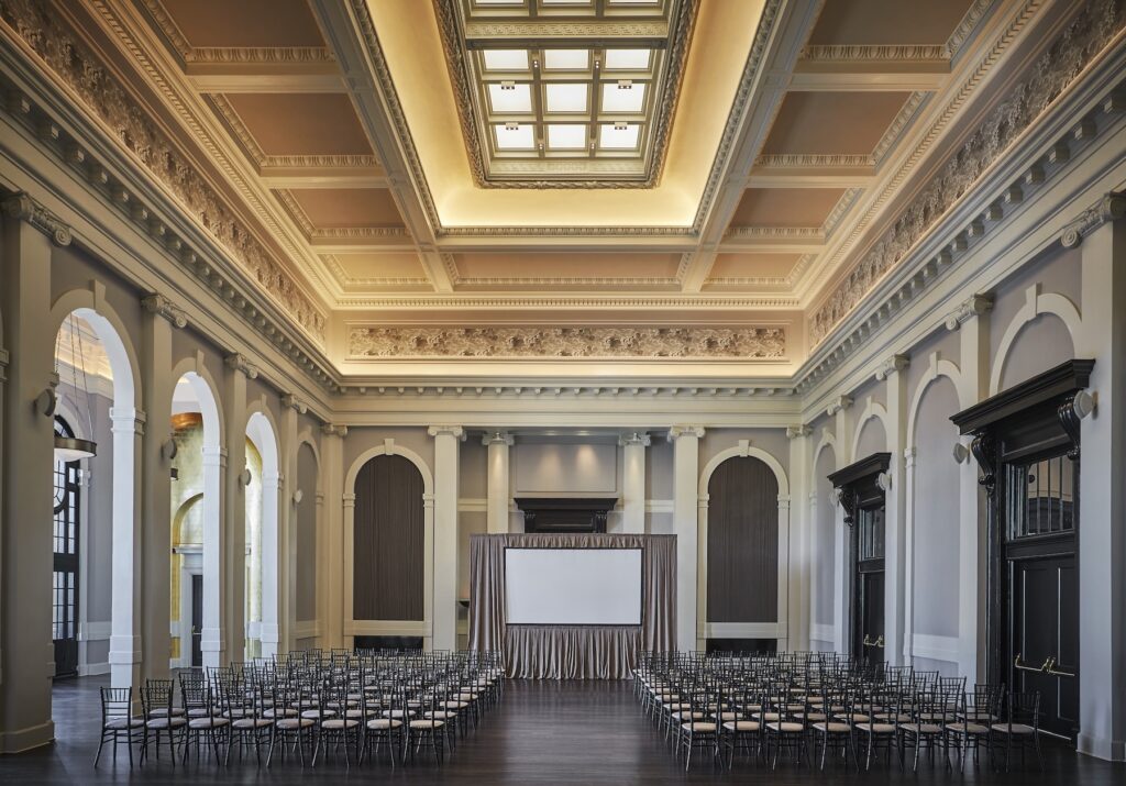 Ballroom with ornate ceiling, arched walls, rows of chairs, and a stage with a projection screen
