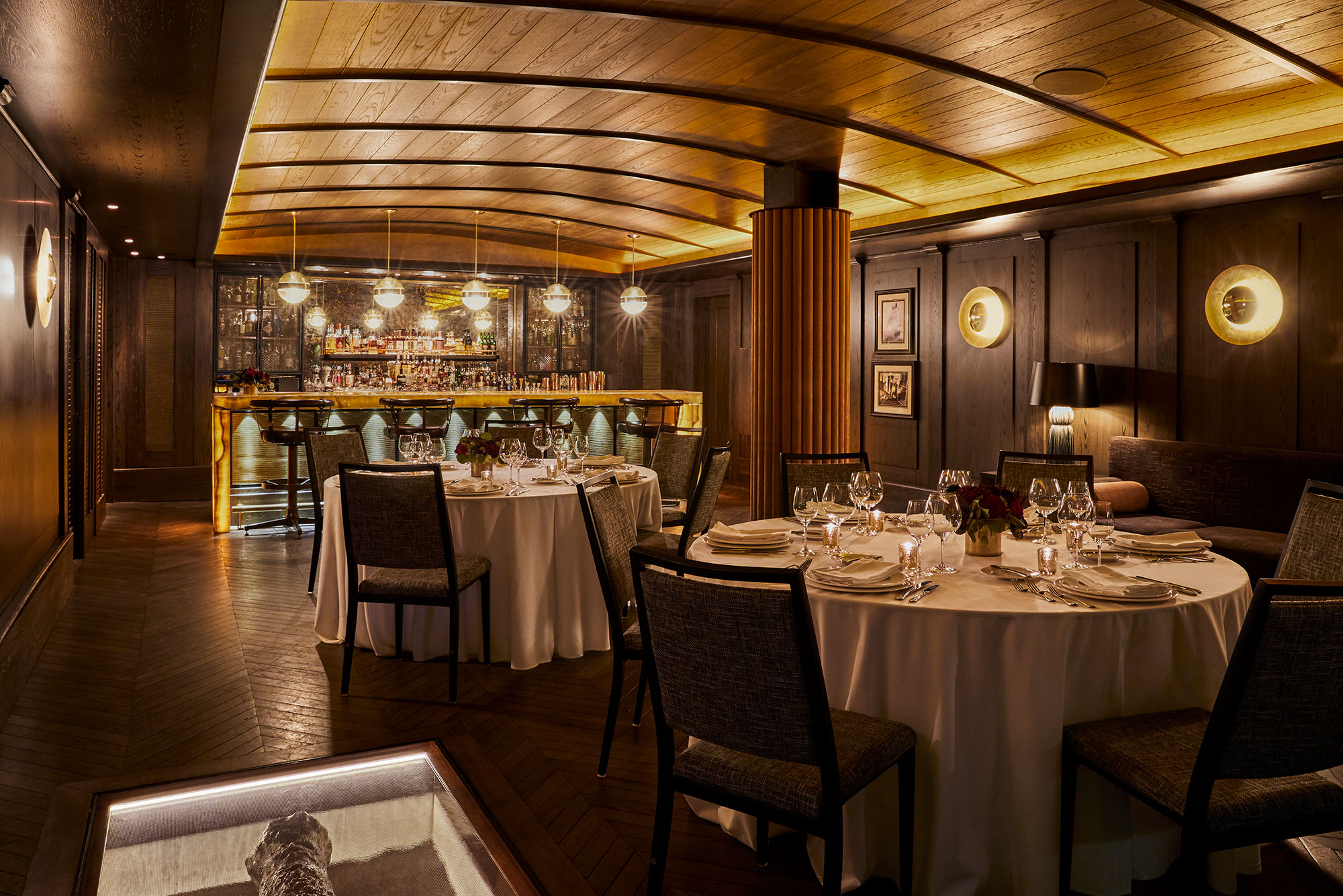 The Cannon Room dining room with round tables, set for dinner beside a warm bar and wood-paneled walls.