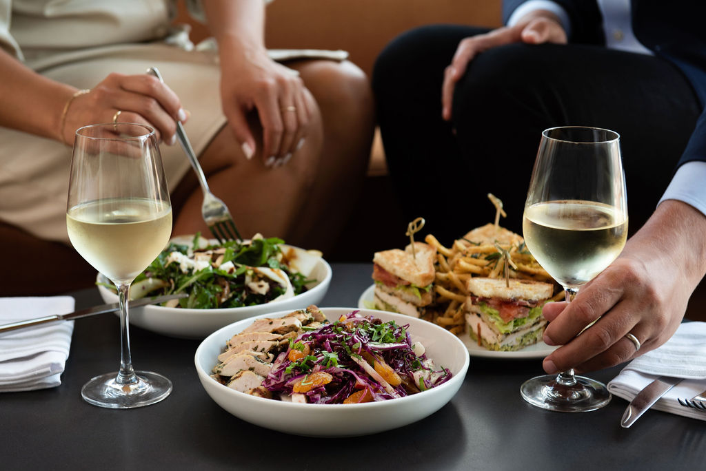 Lunch setting at Pendry Manhattan West with salads, a club sandwich with fries and glasses of white wine.