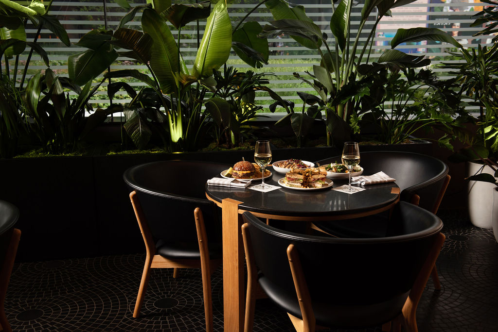 Table setting at the Garden Room with plates of food, glasses of white wine, and lush green plants in the background at Pendry Manhattan West.