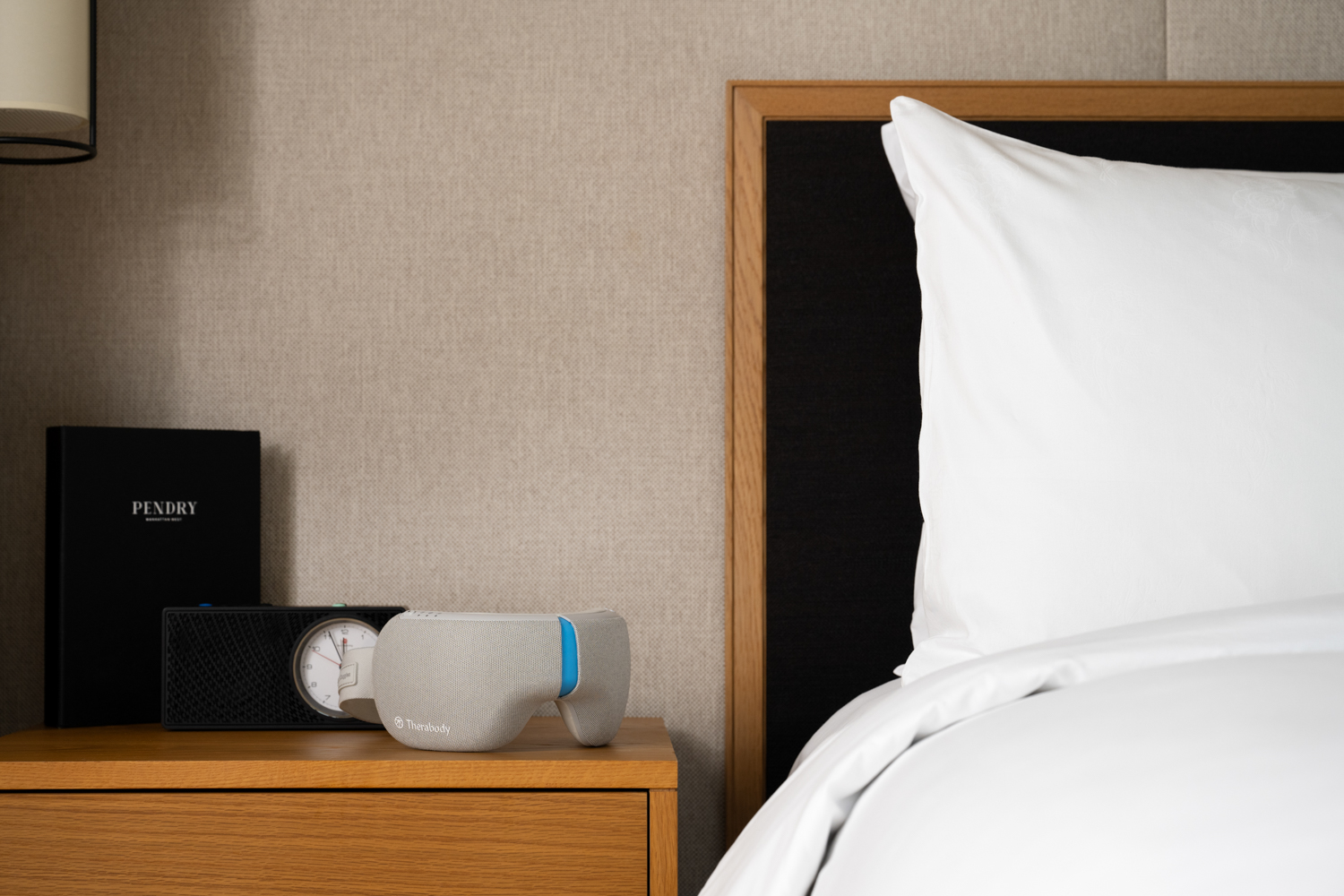Bedside table at Pendry Manhattan West with a Therabody sleep mask, clock, and hotel directory beside a white pillow and bedding.