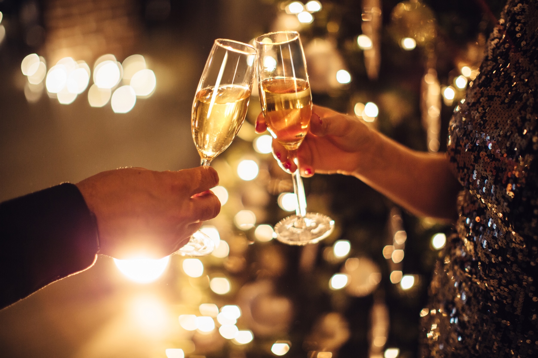 Two people clink champagne glasses in a festive setting, with warm golden lights and holiday decorations in the background