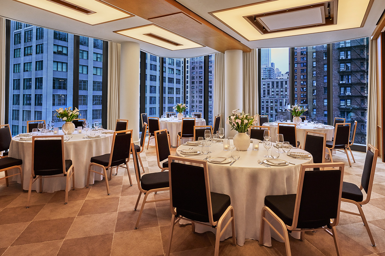 meeting and event space with round tables set for dining, white tablecloths, floral centerpieces, and large floor-to-ceiling windows offering city views