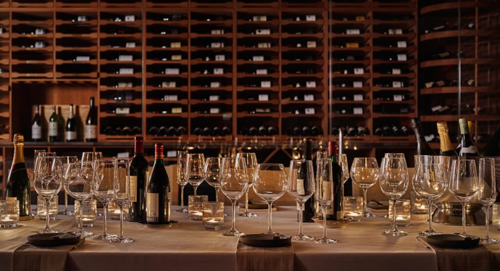 Wine cellar dining setup with bottles, glasses, and candles on a table surrounded by wine racks
