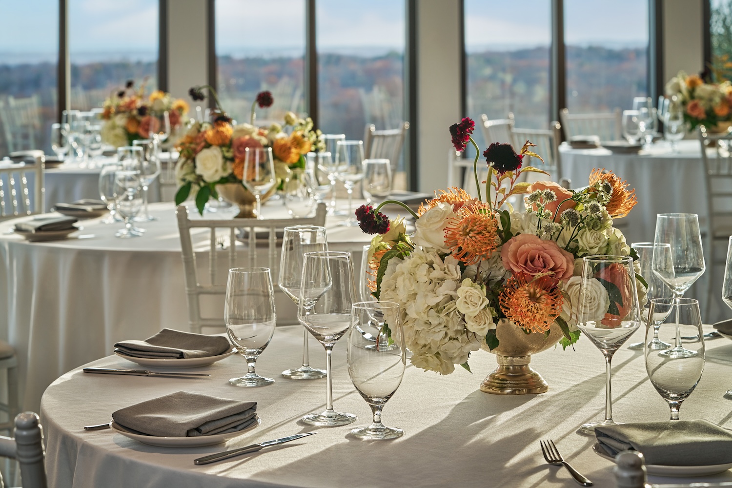 Elegant ballroom setup with round tables draped in white linens, set with glassware and silverware, and decorated with vibrant floral centerpieces featuring roses