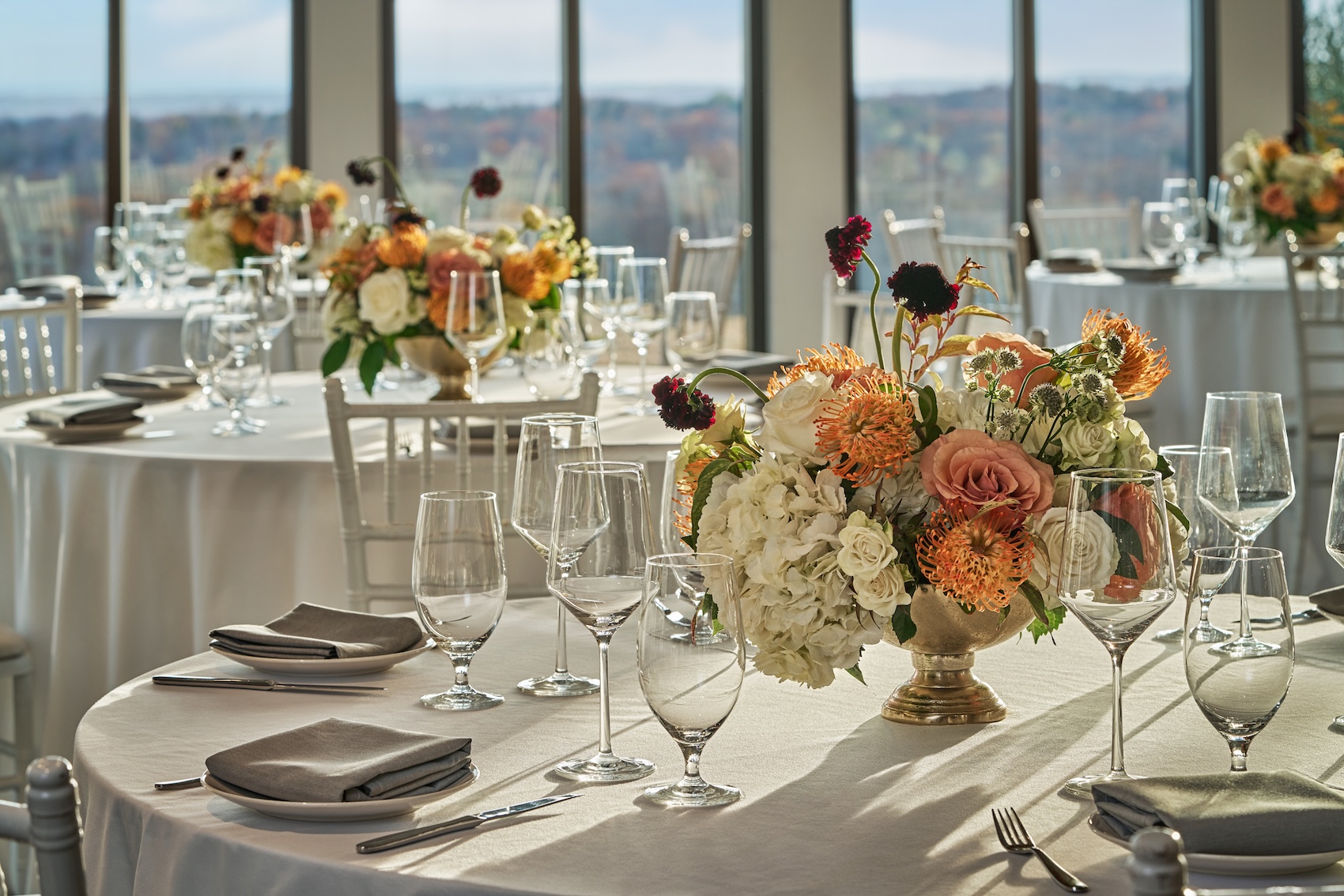 Ballroom is set up for an event with round tables draped in white linens, each adorned with elegant floral centerpieces of roses, hydrangeas, and orange pincushion proteas