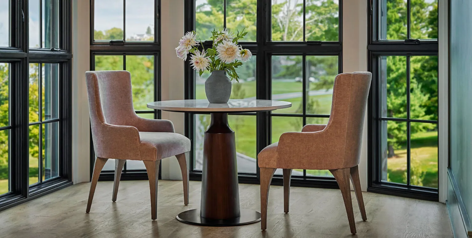 Two pink chairs tucked under a small dining table in front of black-framed windows at Pendry Natirar