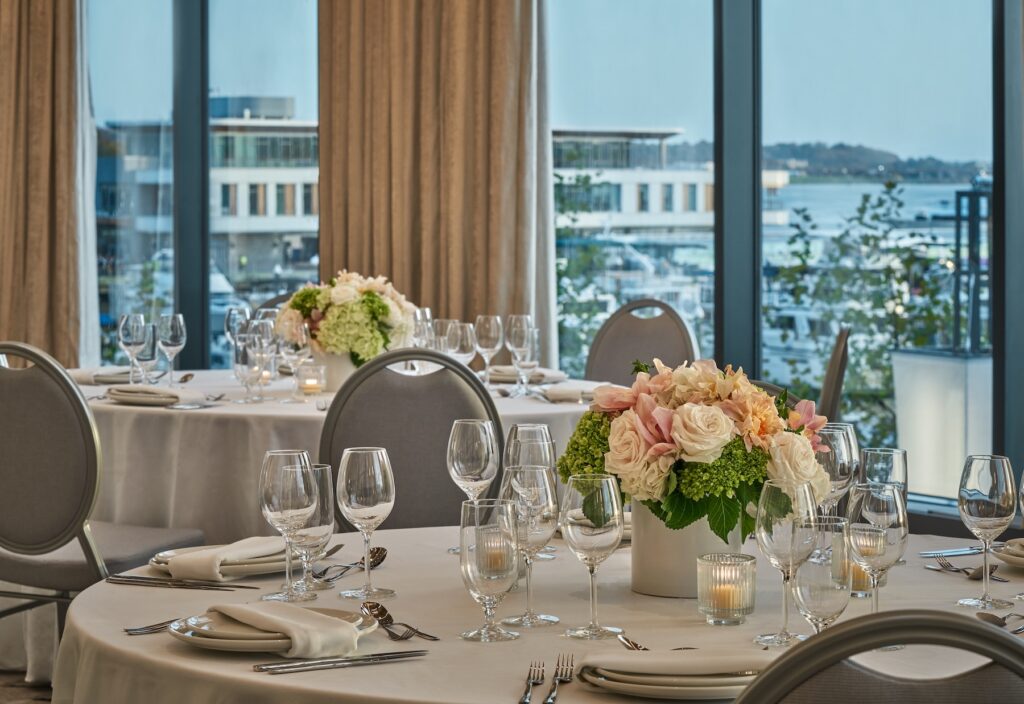 Ballroom setup with round tables, white linens, floral centerpieces of roses and hydrangeas, glassware neatly arranged, and large windows offering marina views