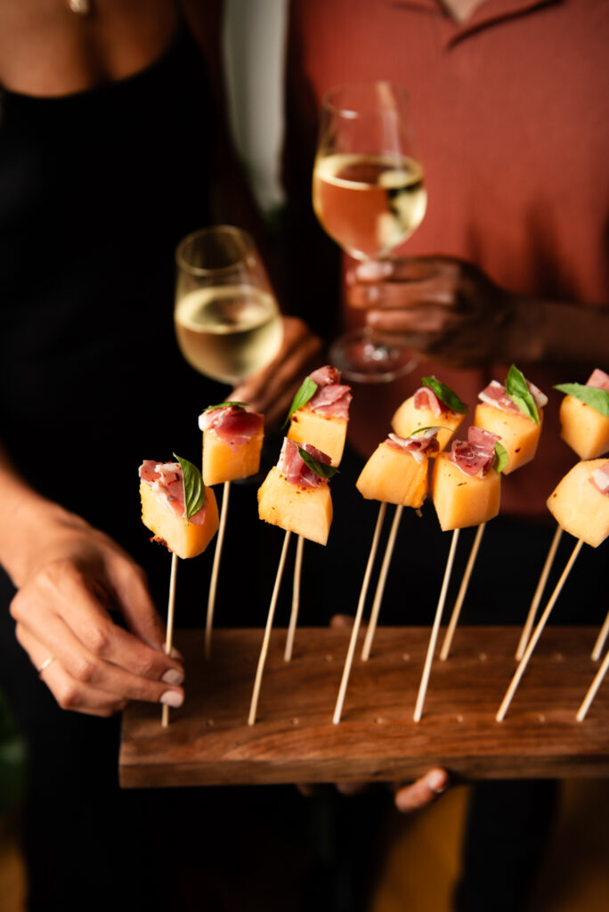 People holding glasses of white wine while serving skewers of melon, prosciutto, and basil on a wooden tray