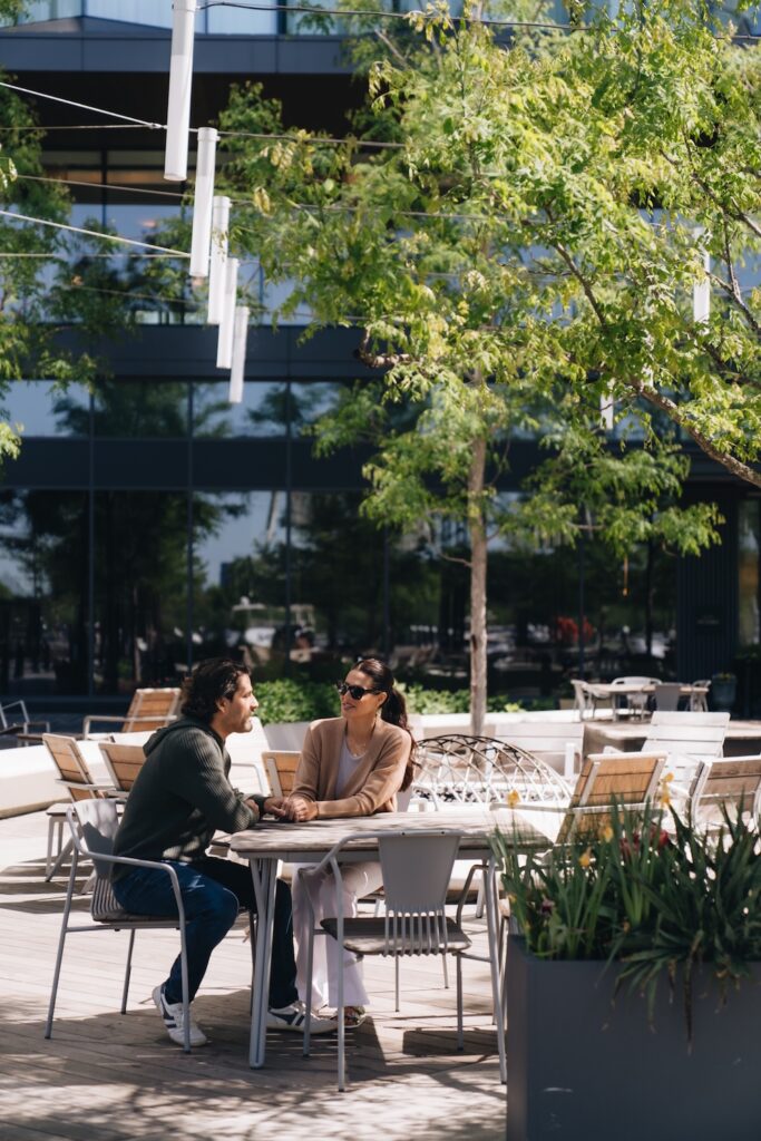 A couple sits outdoors at a wooden table under string lights and trees, holding hands and talking on a sunny day